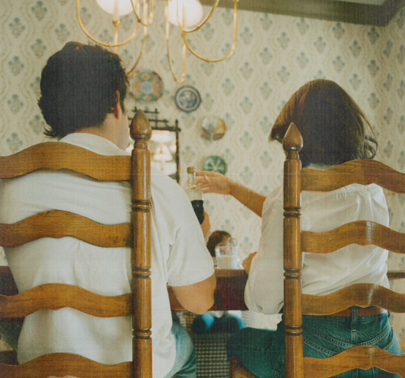 Two people seated at a dining table in ladder-back chairs, in a vintage dining room with patterned wallpaper